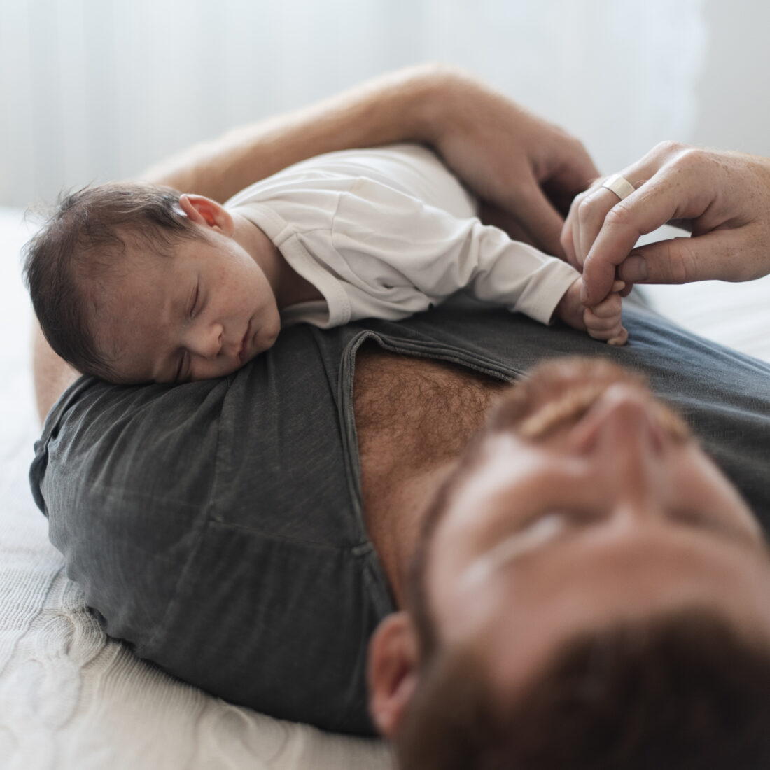 Un neonato dorme appoggiato al petto del papà, che ha gli occhi chiusi e una maglietta grigia