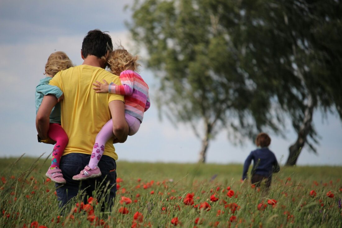 Un papà visto di spalle con due bambini in braccio cammina per un campo di papaveri in una giornata di sole
